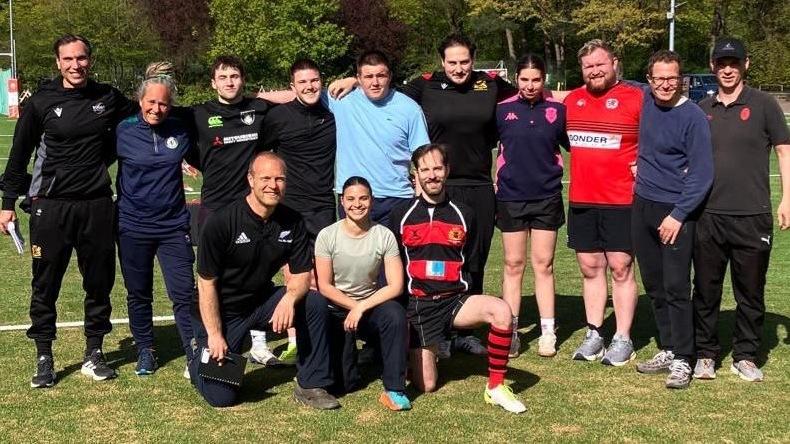 Eine Gruppe von 13 m&auml;nnlichen und weiblichen Trainern und Trainerinnen bei Sonneschein auf dem Kunstrasenplatz der Rugby-Arena Stadtpark