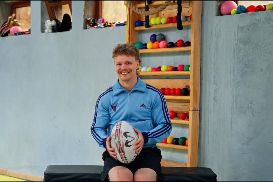 Athletiktrainer Niels Nürnberger sitzt vor einem Regal mit Bällen und hält einen Rugbyball in der Hand